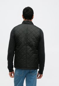 Czarna pikowana kamizelka z wysokim kołnierzem, zestawiona z fakturowanym szarym swetrem, noszona na niebieskich dżinsach, widziana od tyłu.