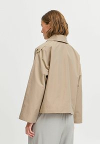 Veste beige courte en matériau léger, dotée d'un col et de détails boutonnés, avec de longues manches larges et une texture lisse.