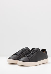 Black leather sneakers with flat laces, rounded toe, and a textured rubber sole. Features padded collar and embossed logo detail.