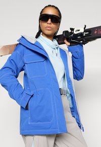 Μπλε ιαματική ζακέτα με γκρι λεπτομέρειες, πολλές μπροστινές τσέπες, κλείσιμο με φερμουάρ, ρυθμιζόμενη κουκούλα, και φορεμένη με μπλε βάση και εξοπλισμό σκι.