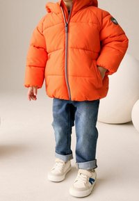 Doudoune orange vif avec capuche, fermeture éclair à l'avant et poches latérales, associée à un jean bleu et des baskets blanches avec des accents bleus.