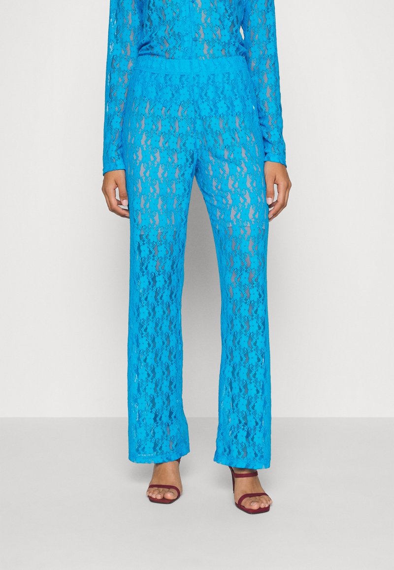 Femme portant un pantalon en dentelle bleu vif et transparent avec un haut assorti, et des talons ouverts bordeaux, debout devant un fond clair uni.