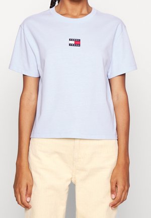 Osoba ubrana w jasnoniebieski krótki t-shirt Tommy Jeans i jasnobeżowe sztruksowe spodnie, stoi z ramionami swobodnie opuszczonymi wzdłuż ciała.