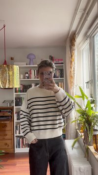 Maglione bianco con strisce blu navy, polsini e orlo a costine. Abbinato a pantaloni neri. Ambientazione interna con librerie e piante sullo sfondo.