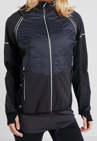 Femme portant une veste de sport zippée noire et gris foncé avec un devant matelassé sur le haut et des passages pour les pouces dans les manches.