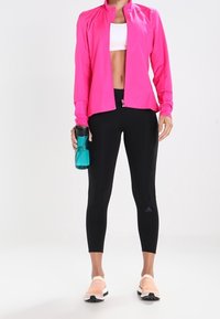 Veste de sport rose, crop top blanc, leggings noirs avec détails en maille, et baskets pêches. Tenant une bouteille d'eau bleue.