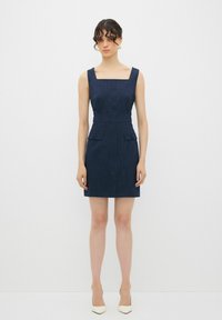 Marineblaues Denimkleid mit quadratischem Ausschnitt, strukturiertem Oberteil und zwei Fronttaschen. Kurze Saumlänge und taillierte Passform, kombiniert mit spitzen weißen Absätzen.