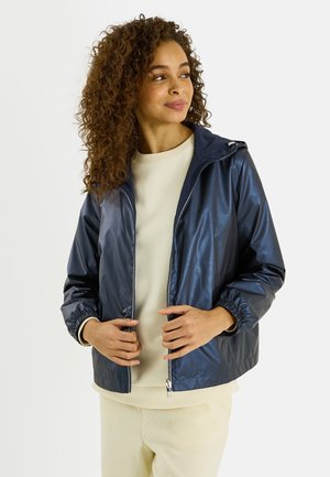 Mujer con cabello rizado que lleva una chaqueta impermeable con capucha azul marino sobre un conjunto color crema, sosteniendo los bordes de la chaqueta y mirando hacia un lado.