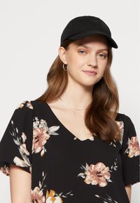 Boné preto usado com uma blusa preta estampada com flores em tons de rosa e creme, com decote em V e mangas curtas. Duas colares prateados em camadas visíveis.