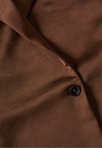 Hnědá tkanina s hladkým povrchem, s černým knoflíkem se čtyřmi dírkami, částečně přehozená a zmačkaná.