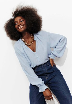 Femme souriante avec un afro volumineux porte un chemisier bleu clair, un jean taille haute foncé, ainsi qu'un collier et un bracelet en chaîne dorée épaisse.
