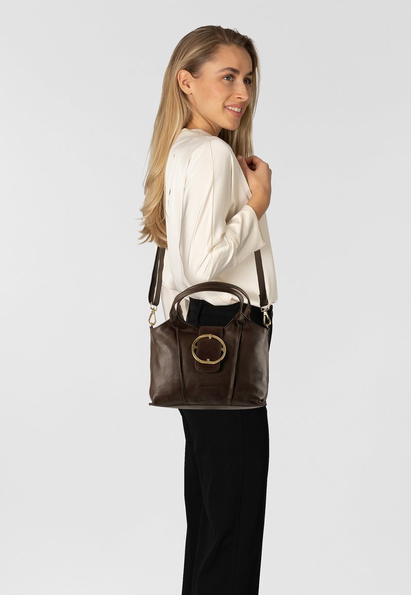 Borsa a mano in pelle marrone con un accento circolare dorato sul fronte, doppi manici e una tracolla staccabile, mostrata su uno sfondo neutro.