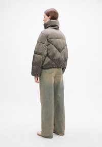 Olivenfarget pufferjakke med høy krage, kviltet design, og en boxy passform, paret med vide, falmede blå jeans og beige sneakers.