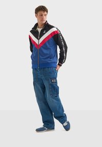 Blå och svart zip-jacka med röd och vit chevron-design. Blå cargobyxor med fickor och logotyp. Blå sportskor.