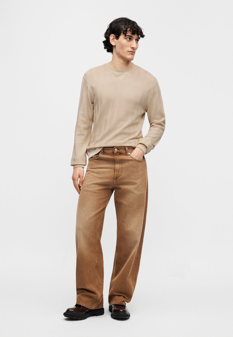 Chemise beige à manches longues avec un motif en chevrons texturé, associée à un jean marron ample et à des bottes noires. Tenue simple et décontractée.