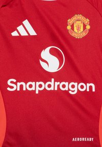 Punane spordijerseys, millel on valged Adidas triibud, Manchester Unitedi emblem ja "Snapdragon" logo. Tehtud tekstureeritud materjalist.