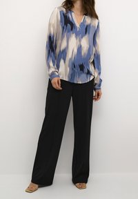 Blouse à motifs bleu et beige avec un col en V, manches longues et coupe décontractée, associée à un pantalon large noir et des sandales beiges.
