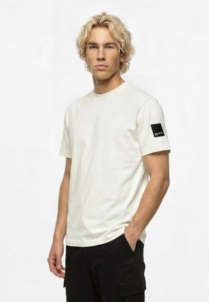 Ung mand med blondt hår iført en ensfarvet hvid kortærmet t-shirt med et sort mærke på venstre ærme og sorte bukser, stående med den ene hånd i lommen.
