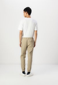 Joggers beige con cinturilla elástica y puños fruncidos, combinados con una camiseta básica blanca de manga corta. Zapatillas blancas visibles en los pies.