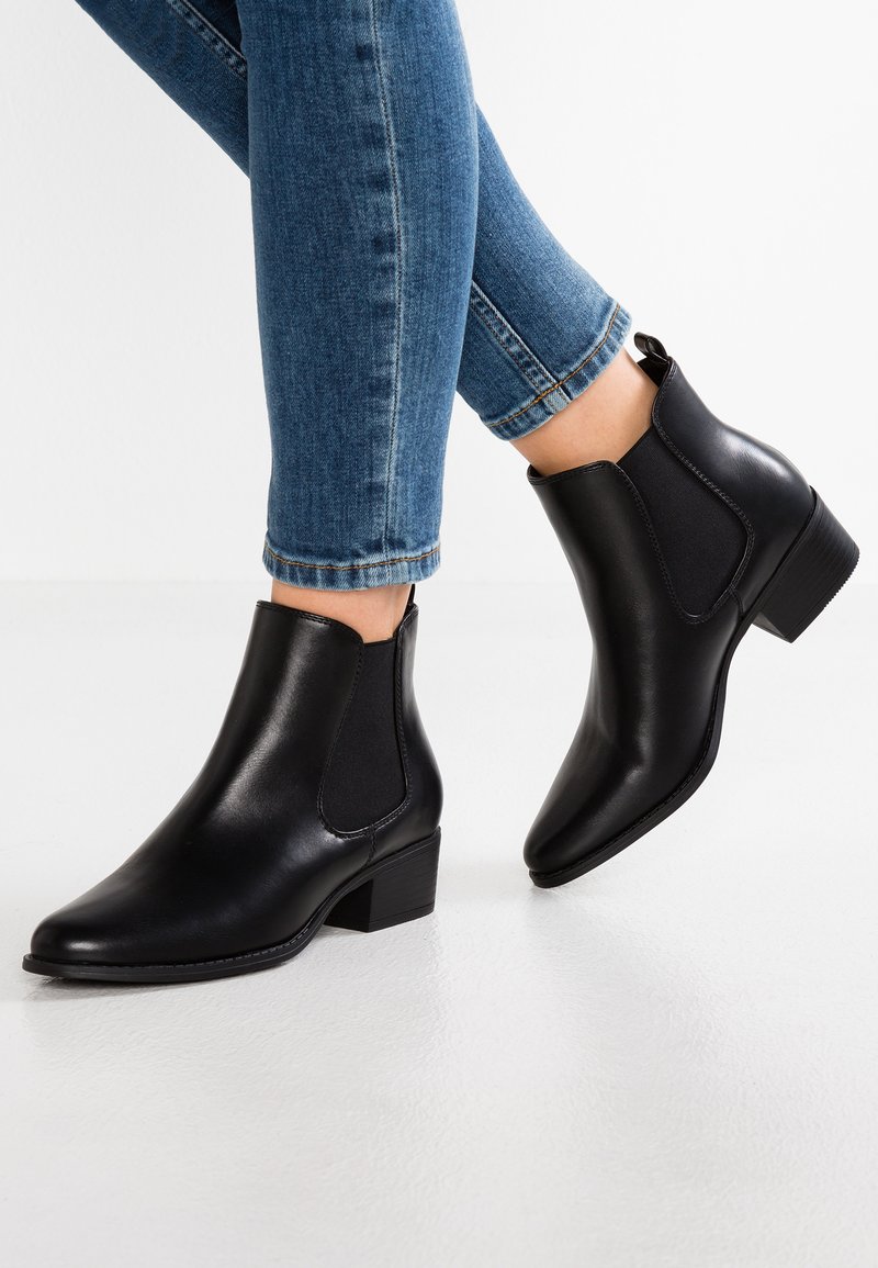 Botas de tobillo de cuero negro con paneles laterales elásticos, punta afilada y un tacón bajo tipo bloque. Conjunto combinado con jeans de mezclilla azul.