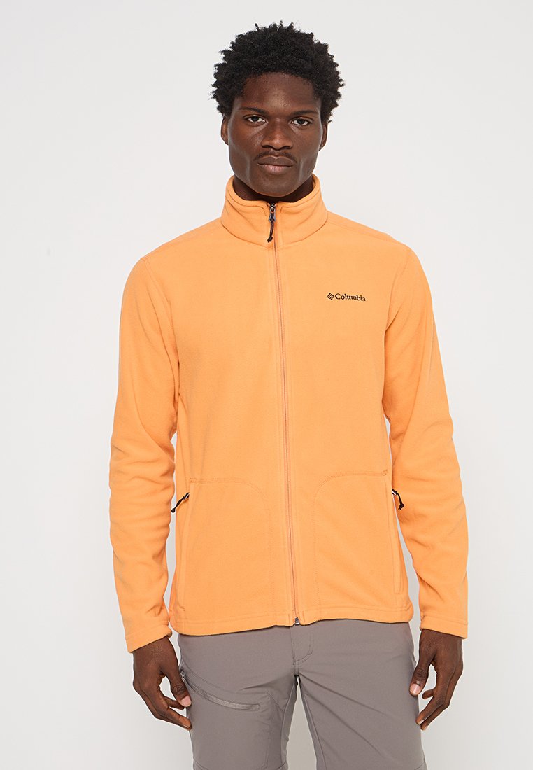 Mężczyzna w jasnopomarańczowej polarowej kurtce Columbia z zapinanym przodem i zapinanymi kieszeniami bocznymi, stojący na jednolitym tle.