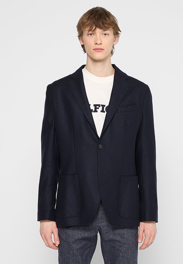 Granatowa wełniana marynarka z wykładanym kołnierzem, zapinana na jeden guzik, z dwiema zewnętrznymi kieszeniami i elegancką sylwetką, która podkreśla dopasowany wygląd.