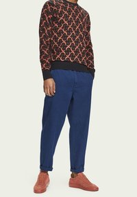 Maglione nero con motivo a onda arancione, pantaloni blu con taglio rilassato e sneakers corallo. Presenta polsini e caviglie a coste, texture morbide.