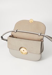 Borsa in pelle scamosciata e cuoio beige con forma arrotondata, hardware circolare in tono oro e tracolla piatta. L'interno presenta una tasca con zip.