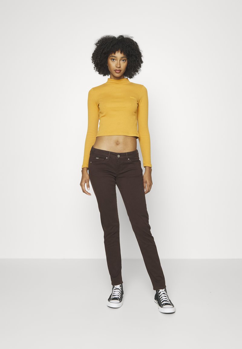 Langärmliges, eng anliegendes, senfgelbes Croptop; braune Skinny Jeans mit Knopfverschluss und Taschen; schwarz-weiße Sneaker.
