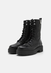 Las botas de combate negras con cordones cuentan con un upper de cuero liso, herrajes plateados, un cierre lateral y una suela de goma negra gruesa.