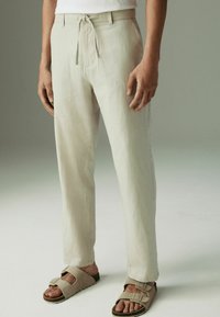 Lichtbeige broek met een trekkoord in de taille, rechte pijpen, zijzakken en een zachte, gestructureerde stof, gedragen met beige sandalen.