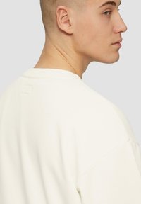 Biała bluza z miękką fakturą, okrągłym dekoltem i opadającymi ramionami. Zawiera małą metkę na karku.