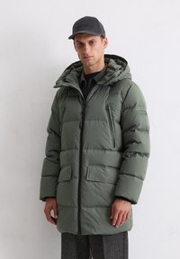 Olive roheline puffjakke kapuutsiga, millel on lukk ja nuppude sulgemine, kaks esimisest taskut ja pehme, tikitud tekstuur. Kantud tumeda särgi kohal.