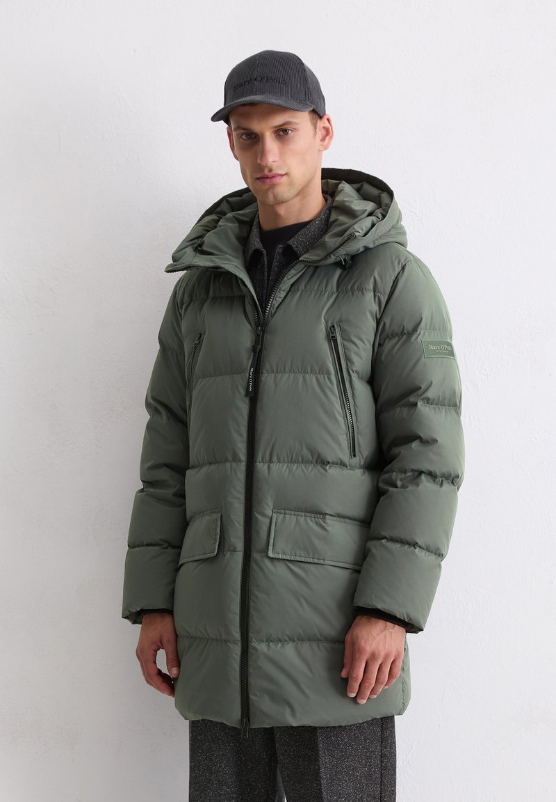 Olive roheline puffjakke kapuutsiga, millel on lukk ja nuppude sulgemine, kaks esimisest taskut ja pehme, tikitud tekstuur. Kantud tumeda särgi kohal.