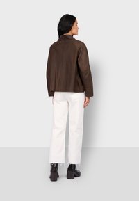 Hnědá kožená bunda s krátkým límcem a spadlými rameny, v kombinaci s bílými širokými kalhotami a černými robustními botami.