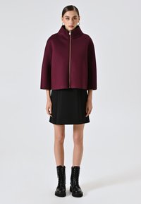 Burgunderfarbener Umhang mit hohem Kragen, goldener Reißverschluss vorne, kombiniert mit einem schwarzen knielangen Kleid und schwarzen Schnürstiefeletten.