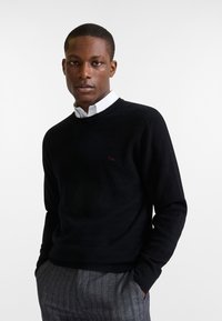 Maglione nero a maglia con un motivo a zigzag testurizzato, scollo a girocollo e un piccolo logo rosso sul petto, abbinato a pantaloni grigi a righe.