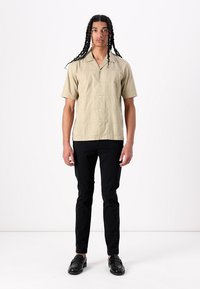 Camisa beige de manga corta en tejido ligero, abotonada, con un collar recto. Pantalones negros ajustados y zapatos negros completan el conjunto.