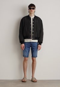 Giacca bomber nera con bottoni a pressione, t-shirt bianca, pantaloni shorts di jeans e sandali beige. Modello con occhiali da sole scuri, design minimalista.