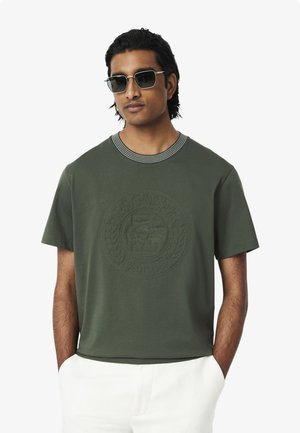 Grünes T-Shirt mit kurzen Ärmeln und einem kreisförmigen, erhabenen Logodesign, ausgestattet mit einem gestreiften Kragen. Aus weichem Stoff, hat einen lockeren Schnitt.