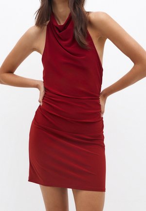 Femme portant une robe rouge ajustée sans manches, avec un col bénitier et une taille froncée, debout les mains sur les hanches devant un fond uni.
