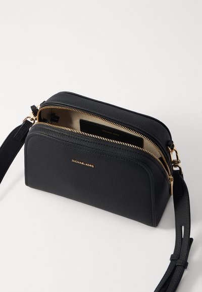 Sac bandoulière en cuir noir avec des zips et des accessoires en or, présentant une texture lisse, un design courbé et un logo sur le devant.