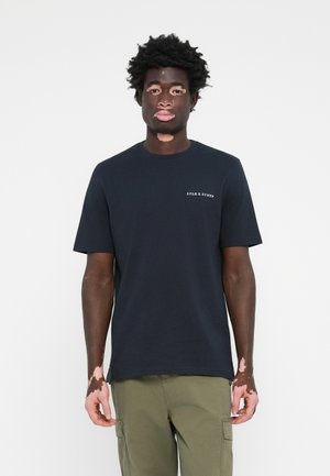 T-shirt en coton noir avec col rond et manches courtes, featuring une broderie de logo blanc sur la poitrine gauche. Le modèle porte un pantalon cargo olive.