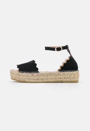 Sort ruskind platformssandal med bølgede kanter, flettet jutesål og ankelrem med guldlås på hvid baggrund.