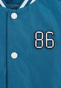 Tyrkysová bomber bunda s bielymi cvokmi a vyšitým číslom "86" v bielej a námorníckej farbe, s textúrou pruhovaného goliera.