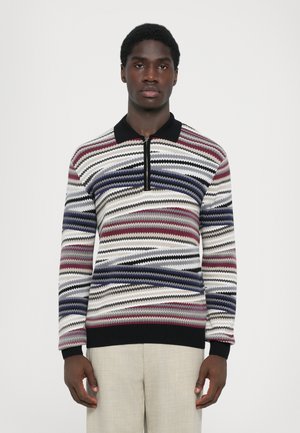 Gestreifter Pullover mit einem Reißverschlusskragen, der Zickzackmuster in Rot, Marineblau, Creme und Schwarz aufweist; gerippter Saum und Ärmelabschlüsse; getragen mit beigen Hosen.