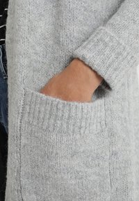 Cárdigan de punto en gris claro con puños acanalados y un gran bolsillo frontal. La textura parece suave y esponjosa, con un ajuste relajado.