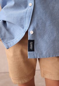 Vaaleansininen napitettava paita, jossa valkoiset napit, yhdistetty vaaleanruskeisiin shortseihin. Paidassa on ommeltu merkintä sivulla brändin tunnistamista varten.