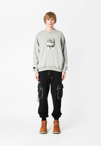 Sudadera gris claro con un estampado gráfico, combinada con pantalones holgados negros que cuentan con bolsillos con cremallera y botas de cordones marrones.