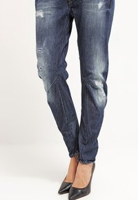 Vaqueros de mezclilla azul oscuro desgastados con un ajuste relajado, que presentan parches desgastados y un ligero desgaste. Combinados con tacones negros de punta puntiaguda.